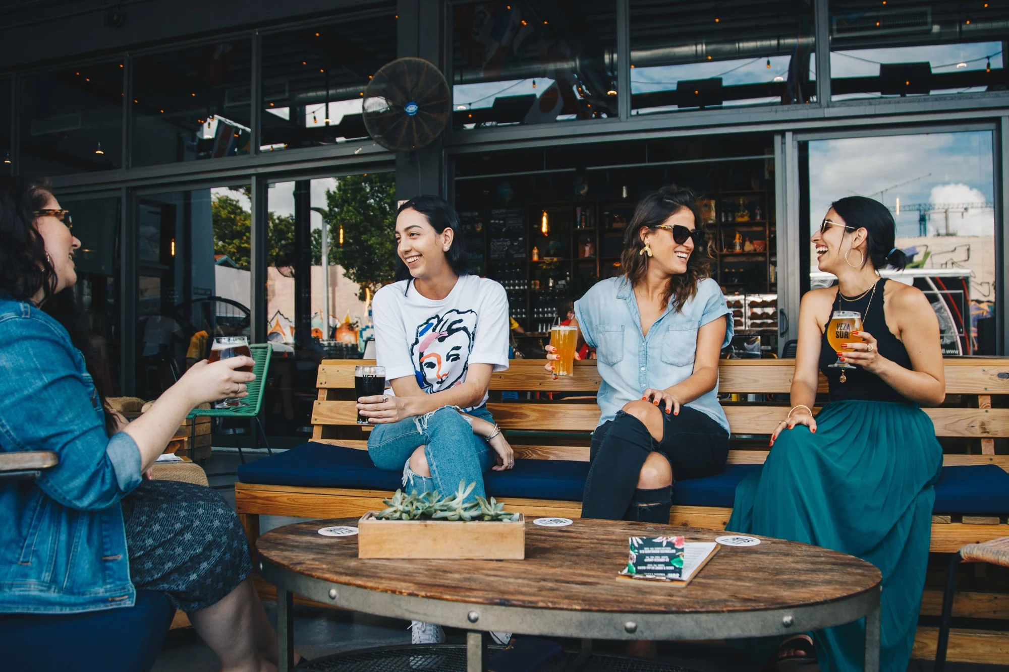 Cuatro mujeres sonríen y conversan mientras sostienen cervezas sentadas en la terraza exterior de un bar.
