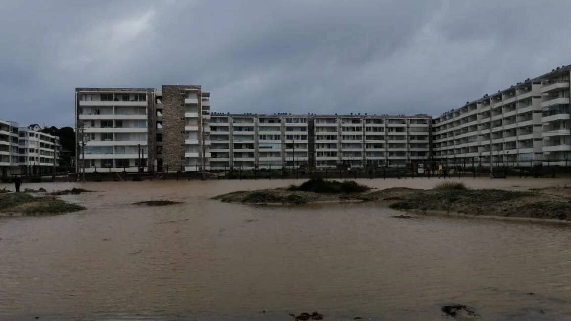 Agua turbia de inundación cubre el área frente a un complejo de edificios de apartamentos bajo cielo nublado.