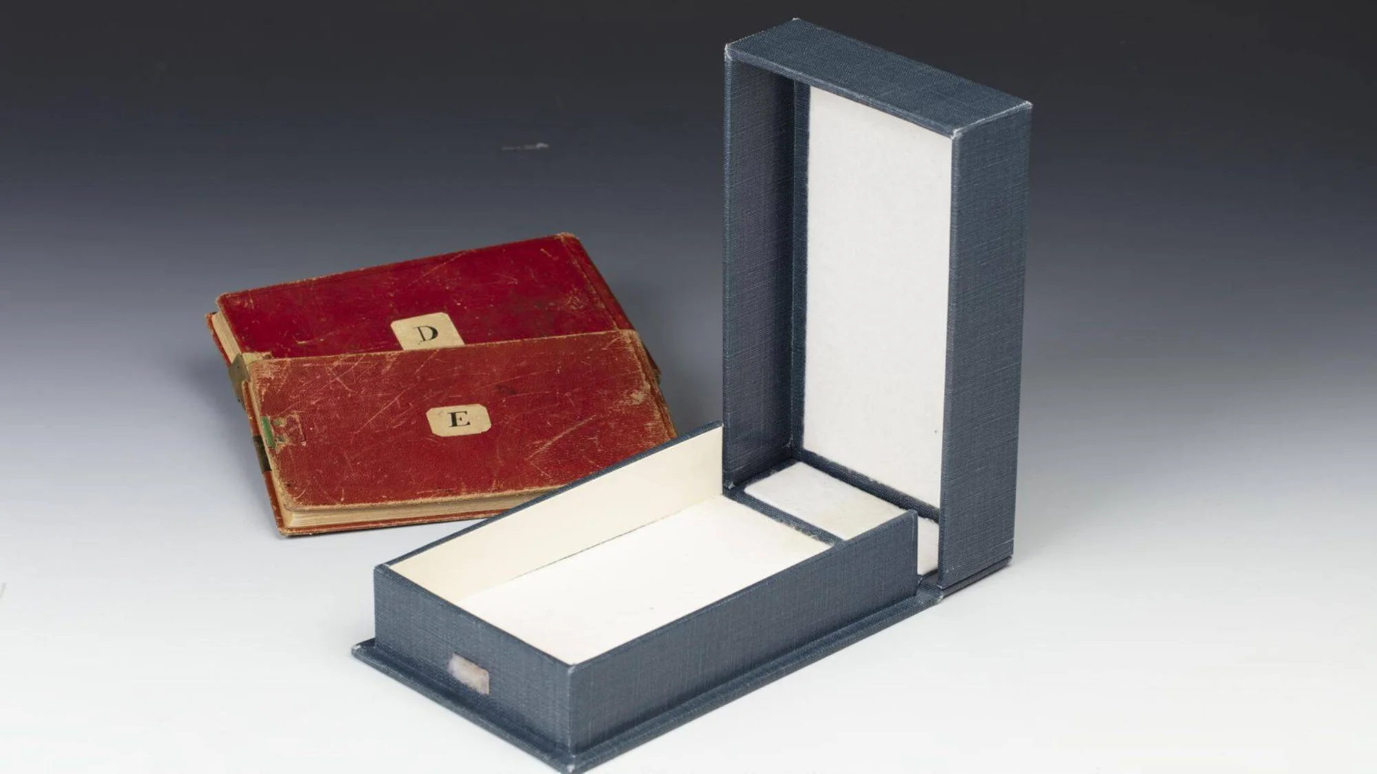 Dos cuadernos rojos desgastados etiquetados con las letras D y E, junto a una caja de archivo azul abierta y vacía.