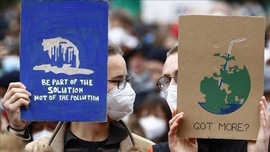 Dos manifestantes con mascarilla sostienen carteles sobre cambio climático: 'Be part of the solution, not of the pollution' y un planeta Tierra con pajita.