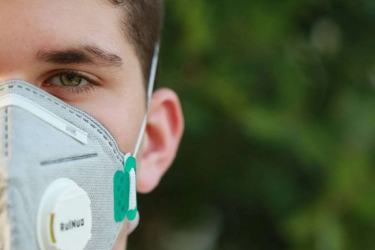 Joven con mascarilla respiratoria con válvula de exhalación frente a vegetación desenfocada.