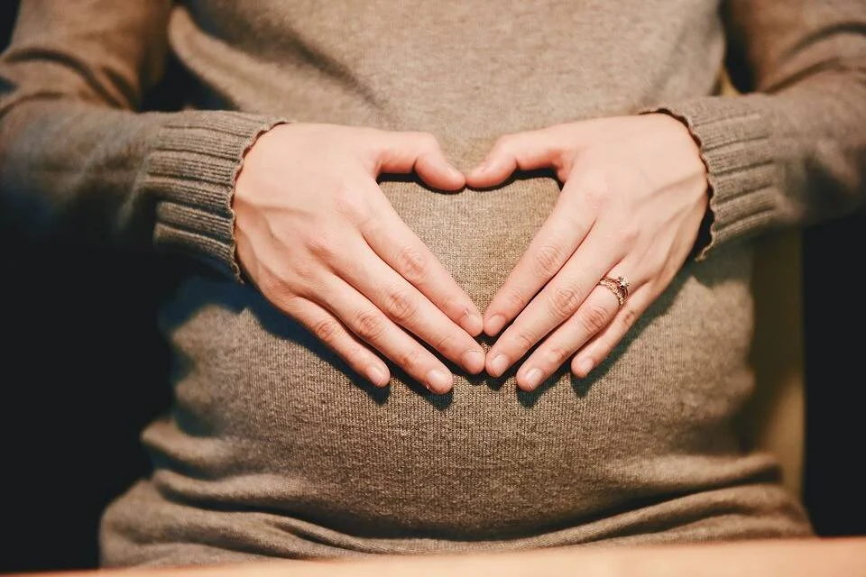 Mujer embarazada forma un corazón con sus manos sobre el vientre, vistiendo un suéter gris.