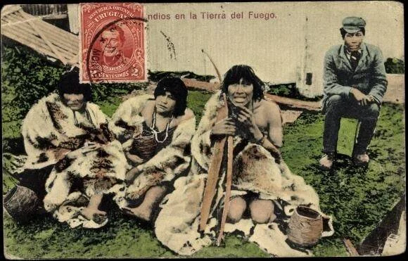 Postal antigua con indígenas de Tierra del Fuego cubiertos con pieles, sentados en el suelo junto a un hombre vestido a la occidental.