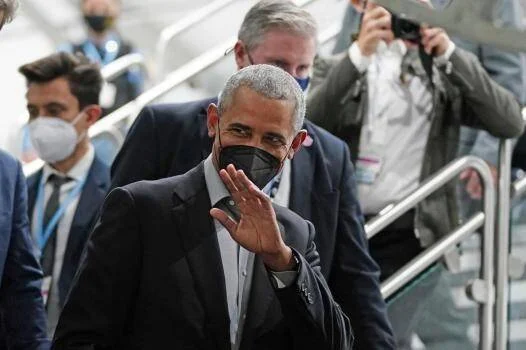Hombre con mascarilla negra saluda con la mano mientras desciende escaleras rodeado de personas en un evento oficial.