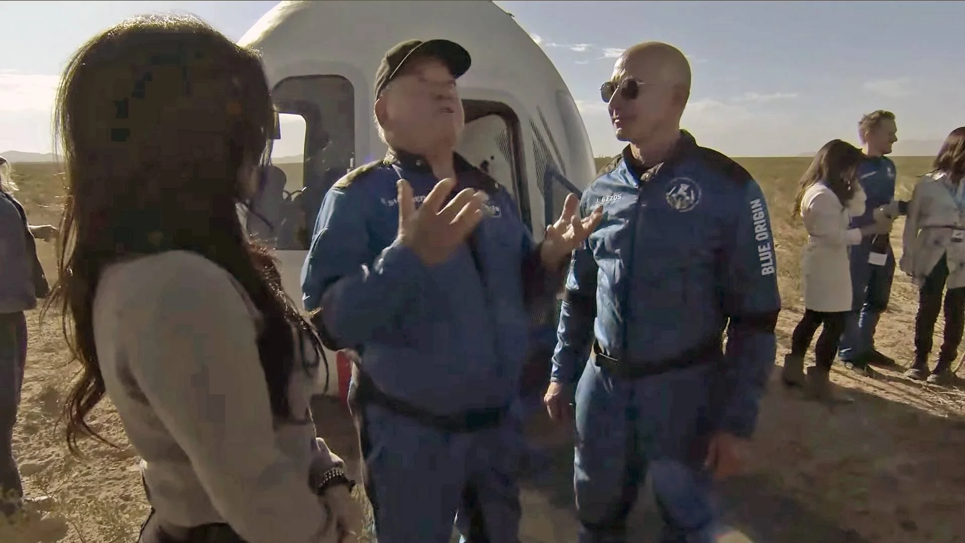 Dos personas con trajes azules de Blue Origin conversan junto a la cápsula espacial New Shepard tras su aterrizaje en el desierto.