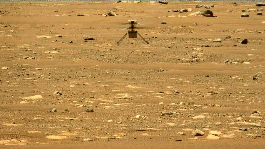 El helicóptero Ingenuity de la NASA eleva vuelo sobre el suelo rojizo y rocoso de Marte.