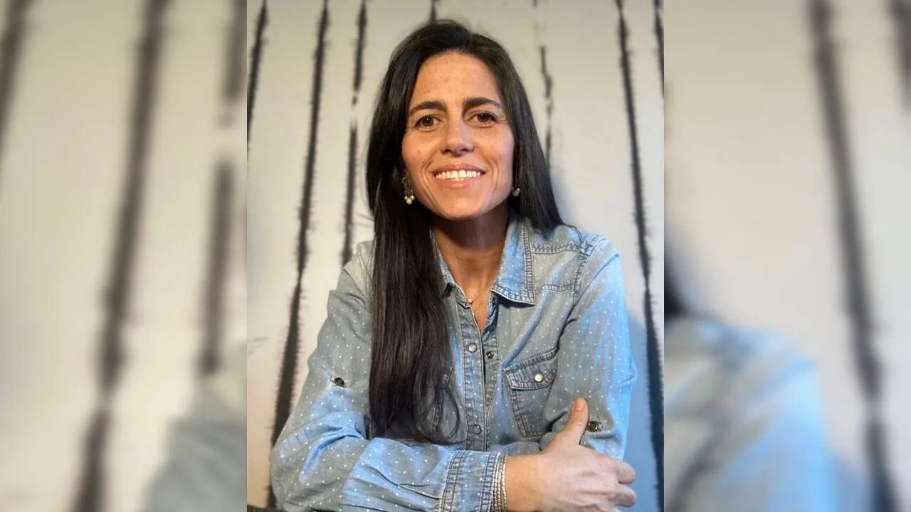 Mujer de cabello largo y oscuro sonríe con los brazos cruzados, vistiendo una camisa de jean celeste.