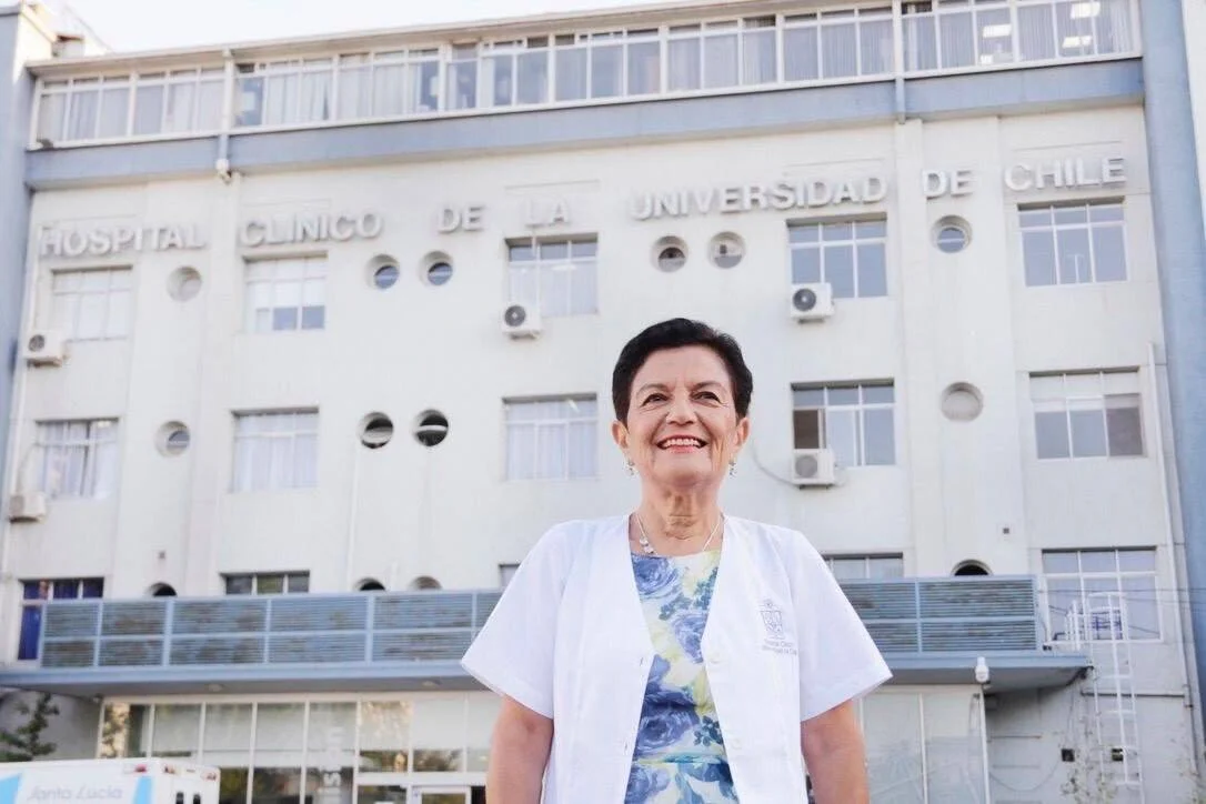Mujer con delantal blanco sonríe frente a la fachada del Hospital Clínico de la Universidad de Chile.