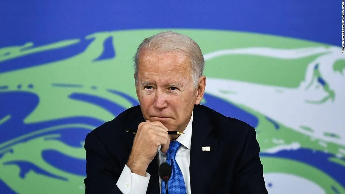 Joe Biden sostiene sus anteojos con gesto pensativo durante una cumbre sobre cambio climático, ante un fondo con motivos ambientales.