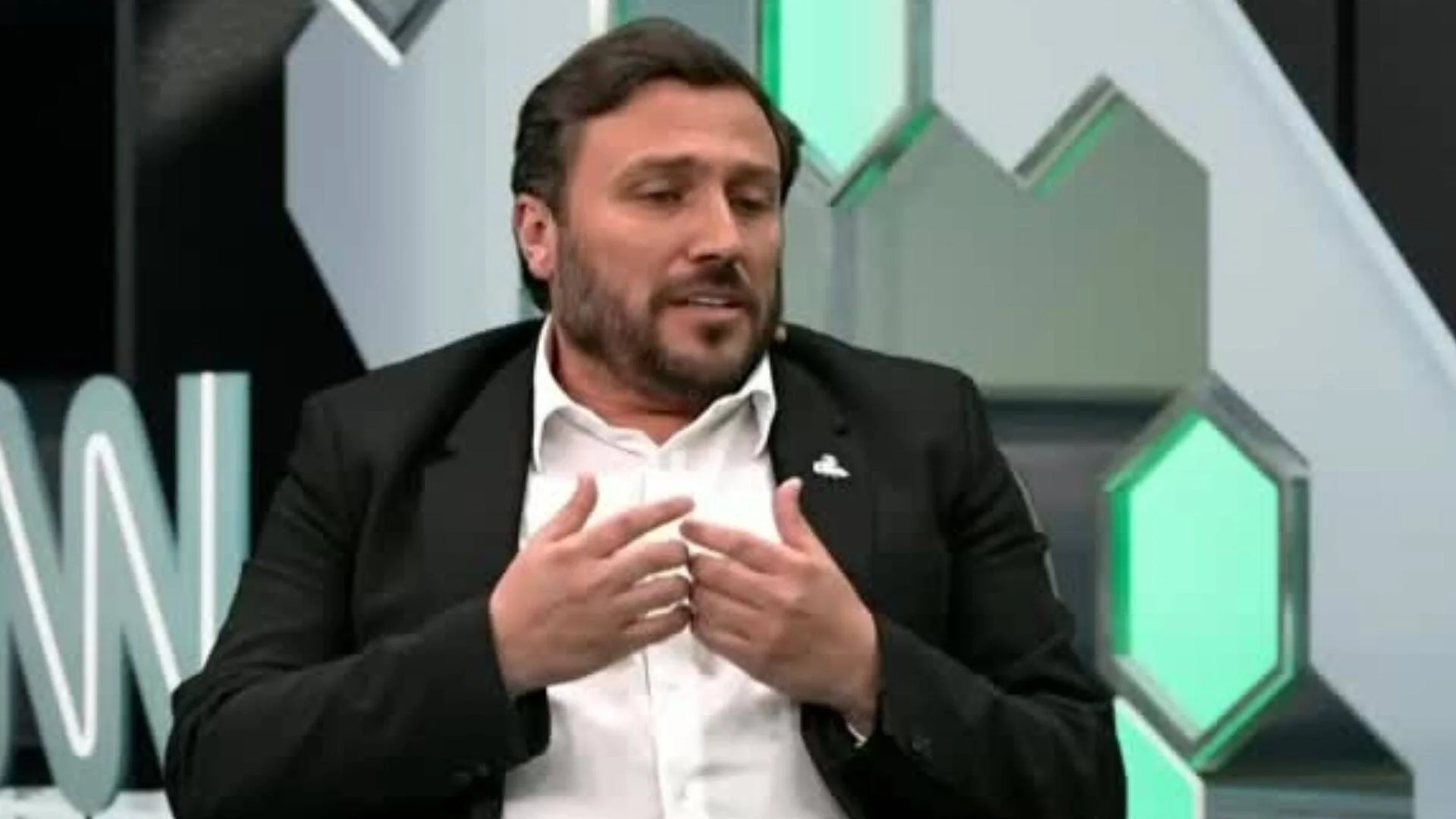 Hombre con traje negro y camisa blanca gesticulando con las manos durante una entrevista en un estudio de televisión.
