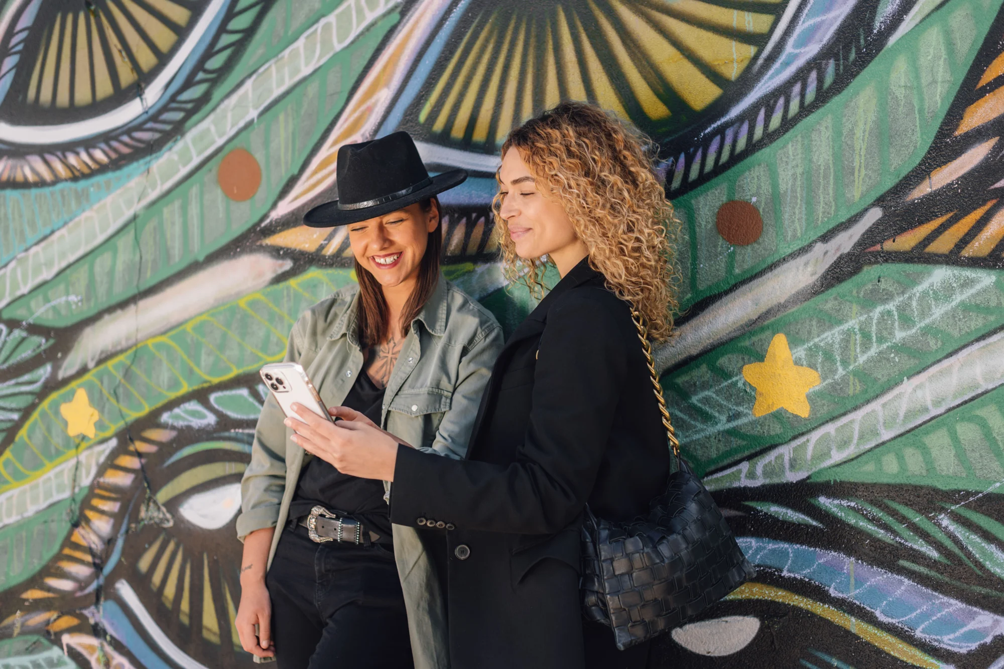 Dos jóvenes sonríen mientras miran juntas la pantalla de un smartphone frente a un colorido mural urbano.