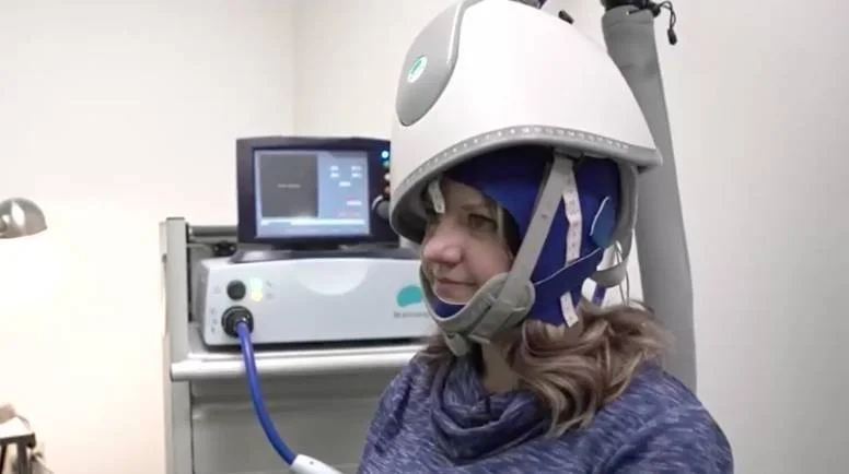 Una mujer lleva puesto un casco de estimulación magnética transcraneal conectado a un equipo médico en un consultorio clínico.
