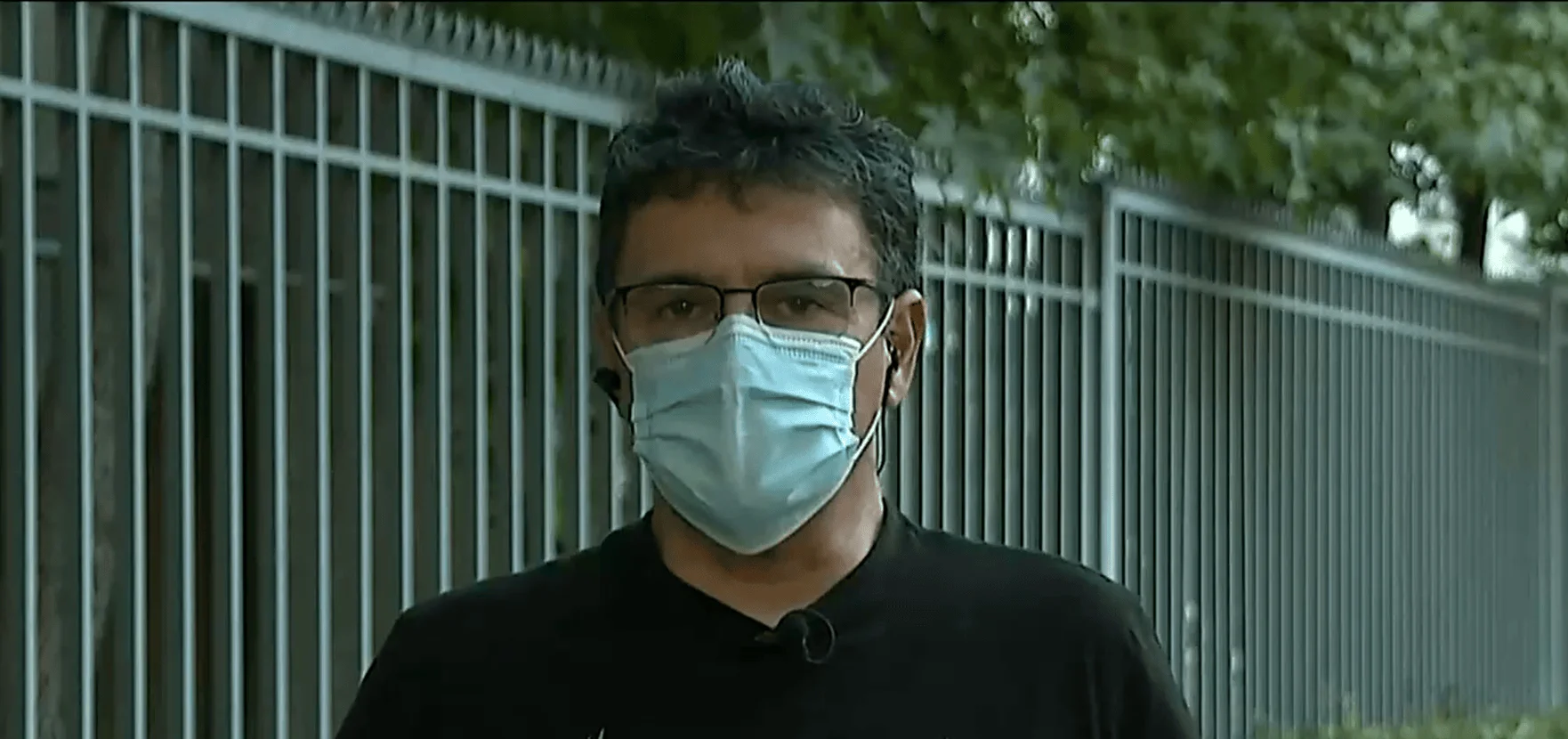 Hombre con gafas y mascarilla quirúrgica azul frente a una reja metálica, con auricular de periodista visible.
