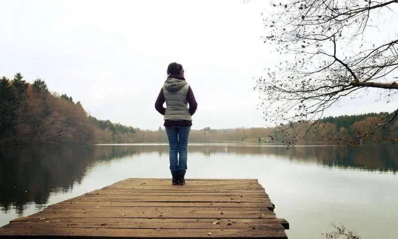 Una mujer de espaldas, de pie en un muelle de madera, contempla un lago tranquilo rodeado de bosque en otoño.