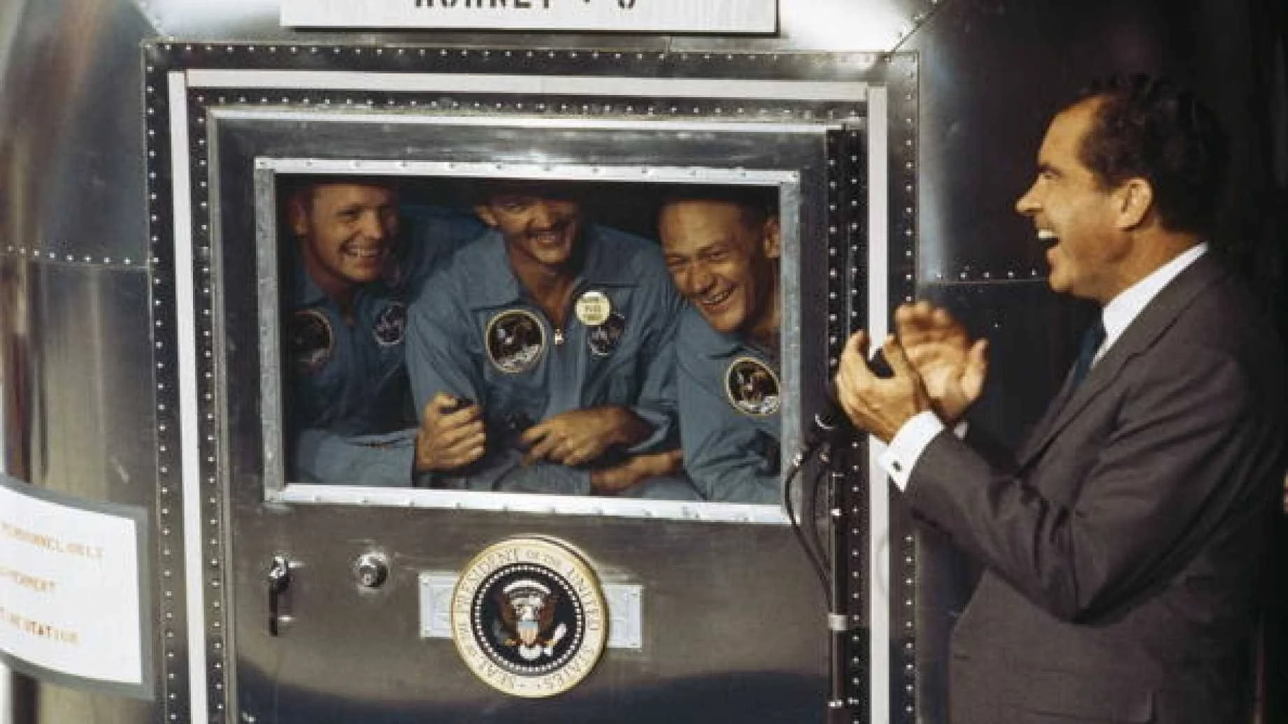 Tres astronautas con trajes azules sonríen desde el interior de un módulo de cuarentena con el sello presidencial, mientras un hombre de traje les saluda aplaudiendo.