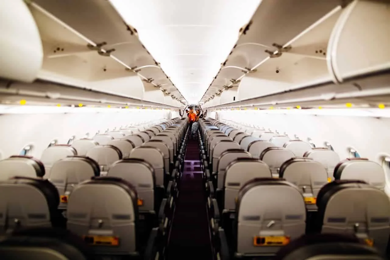 Cabina de un avión comercial vacía con filas de asientos grises y un tripulante de cabina al fondo del pasillo.