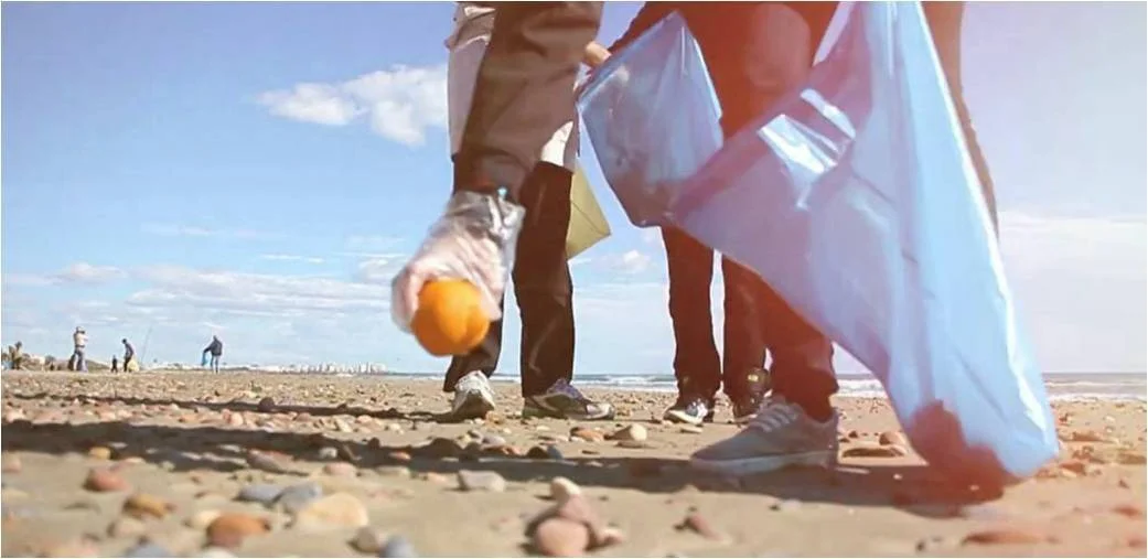 Voluntarios con guantes y bolsas recogen basura en una playa, vistos desde el nivel del suelo.