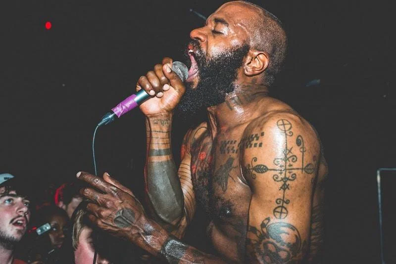 Cantante con el torso descubierto y tatuajes sostiene el micrófono mientras canta apasionadamente ante el público en un concierto.