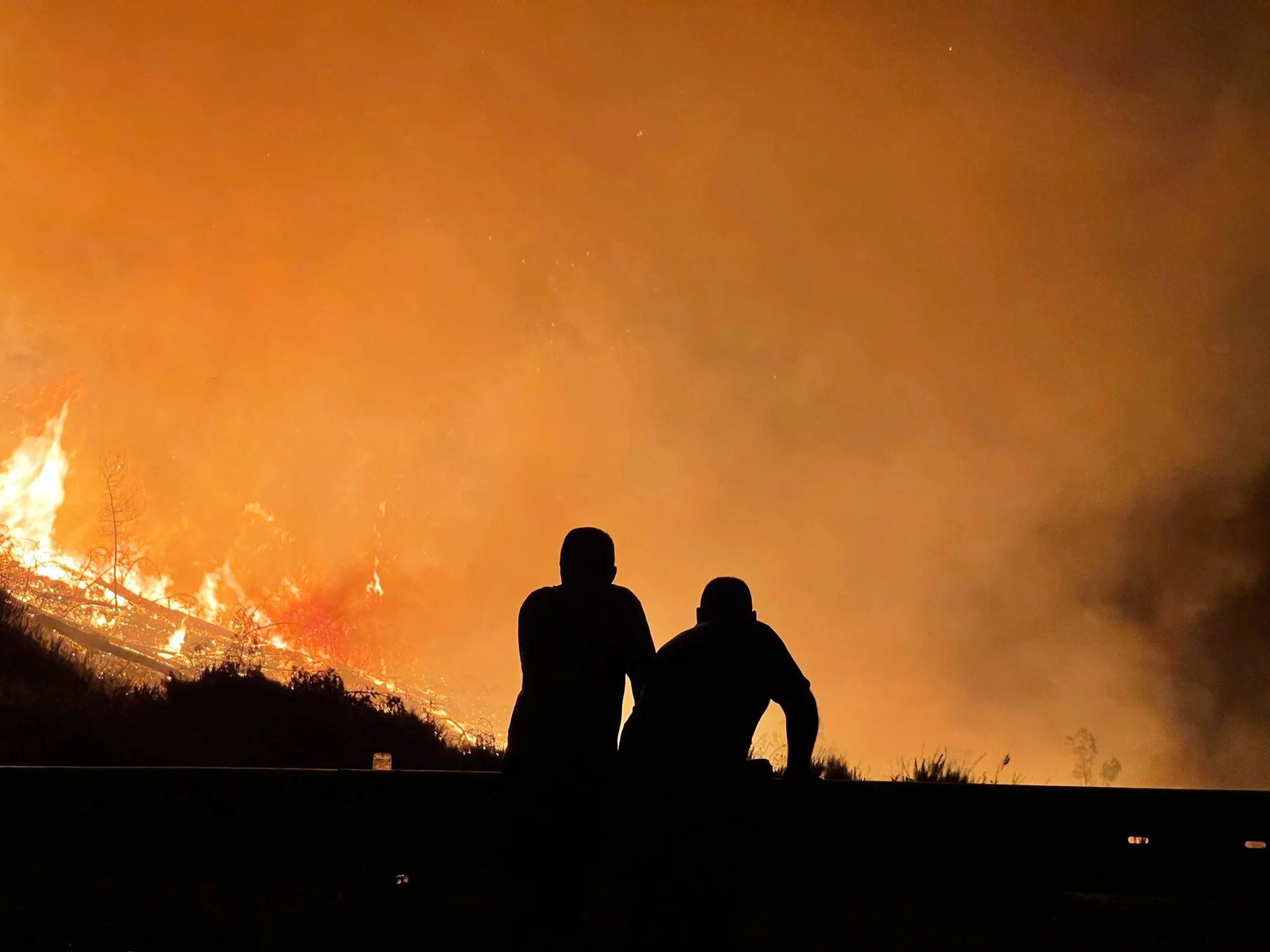 Dos personas en silueta observan un intenso incendio forestal que ilumina el cielo de naranja durante la noche.