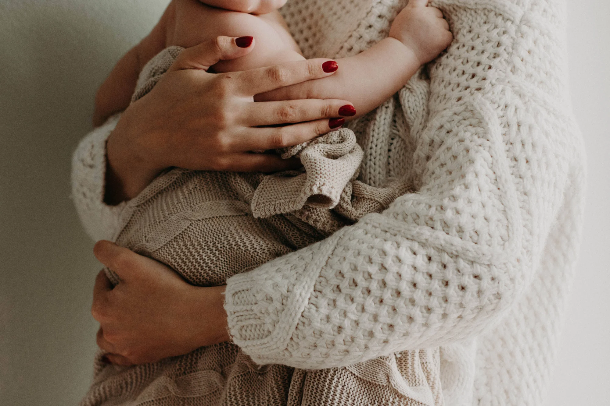 Persona con suéter blanco de punto abraza a un bebé envuelto en manta tejida beige, sujetándolo con ambas manos de uñas rojas.