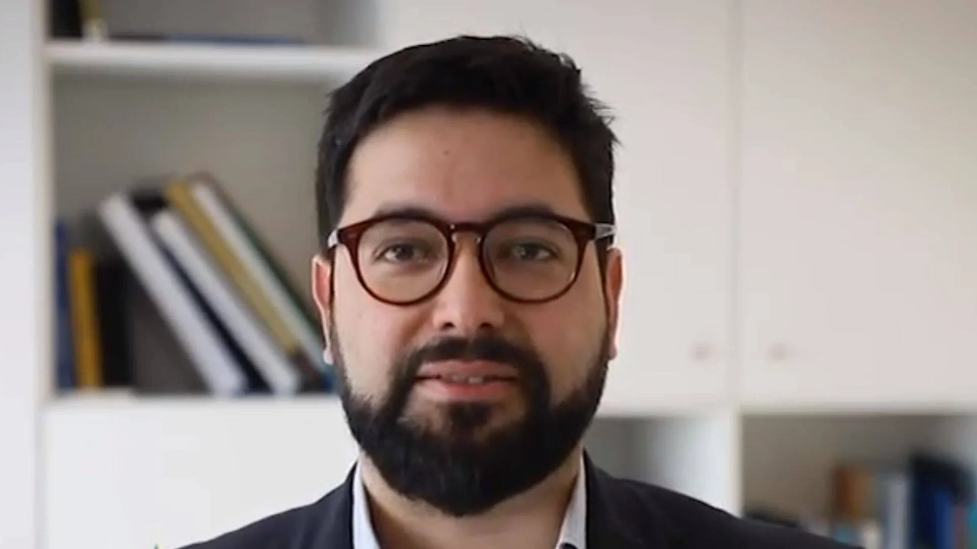 Hombre joven con barba, gafas de montura carey y traje oscuro habla frente a una estantería con carpetas.