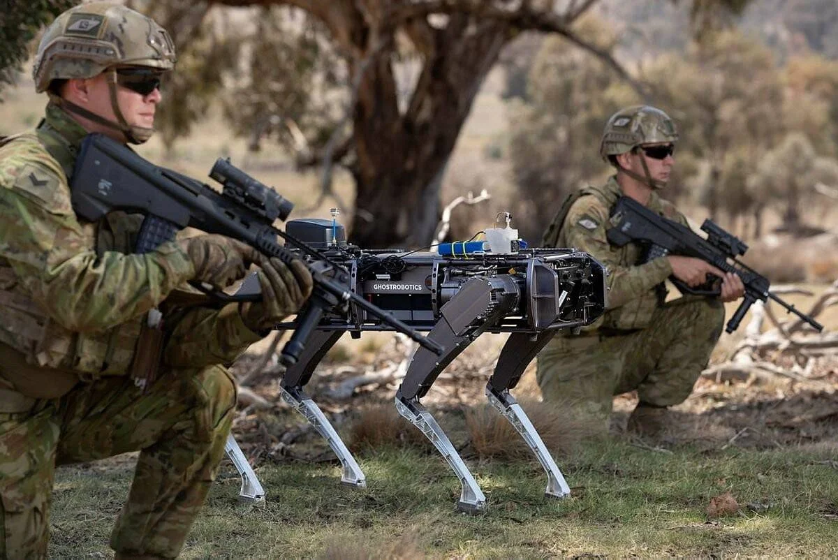 Dos soldados armados en uniforme de campaña flanquean un robot cuadrúpedo de la marca Ghostrobotics durante un ejercicio militar en exterior.