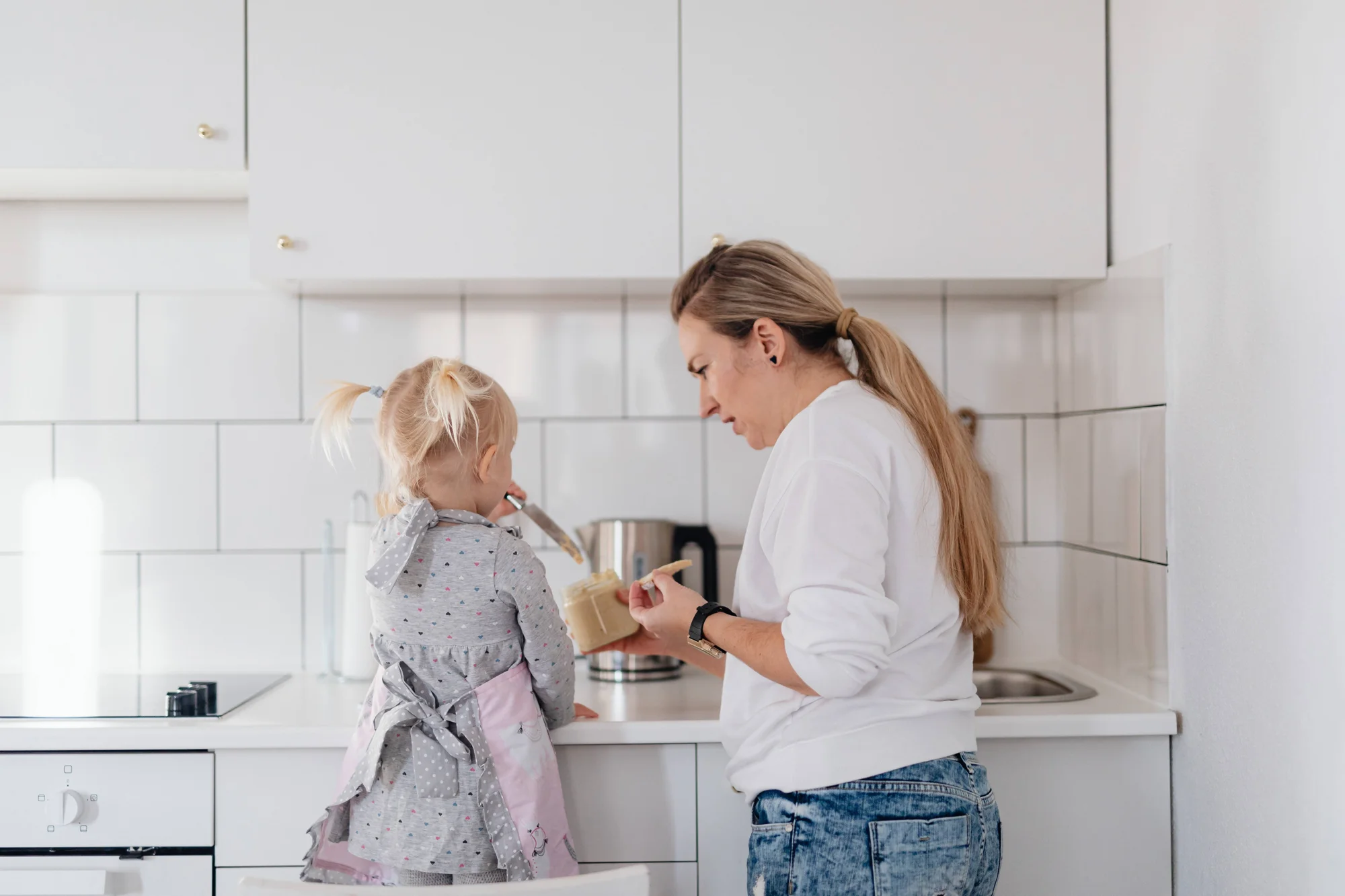 Una mujer le ofrece a una niña pequeña una cucharada de mantequilla de maní en una cocina moderna blanca.