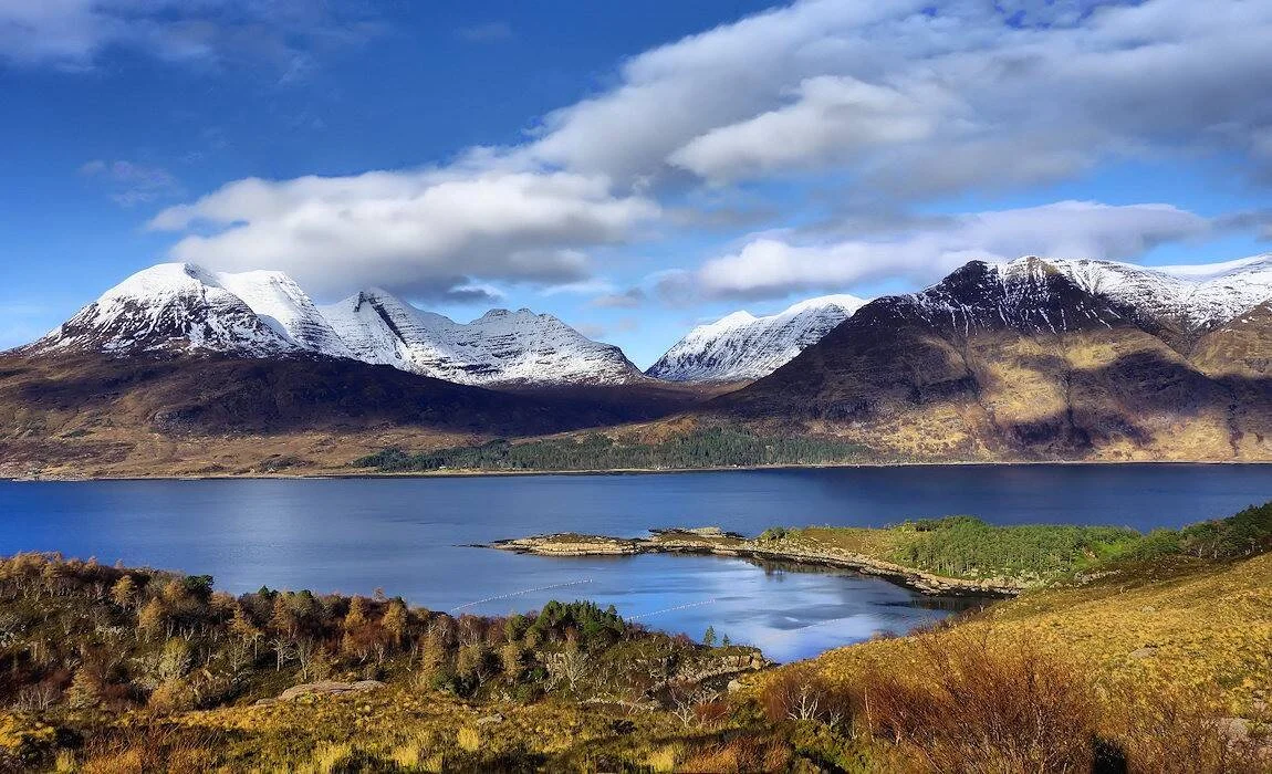 Lago tranquilo rodeado de montañas con cimas nevadas bajo un cielo azul con nubes, paisaje de las Highlands escocesas.