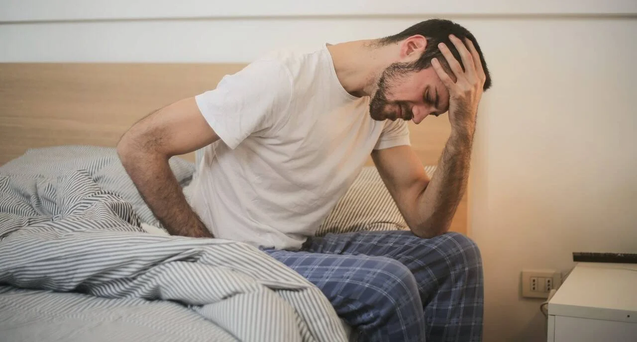 Hombre en pijama sentado al borde de la cama sosteniéndose la cabeza con gesto de dolor o malestar.