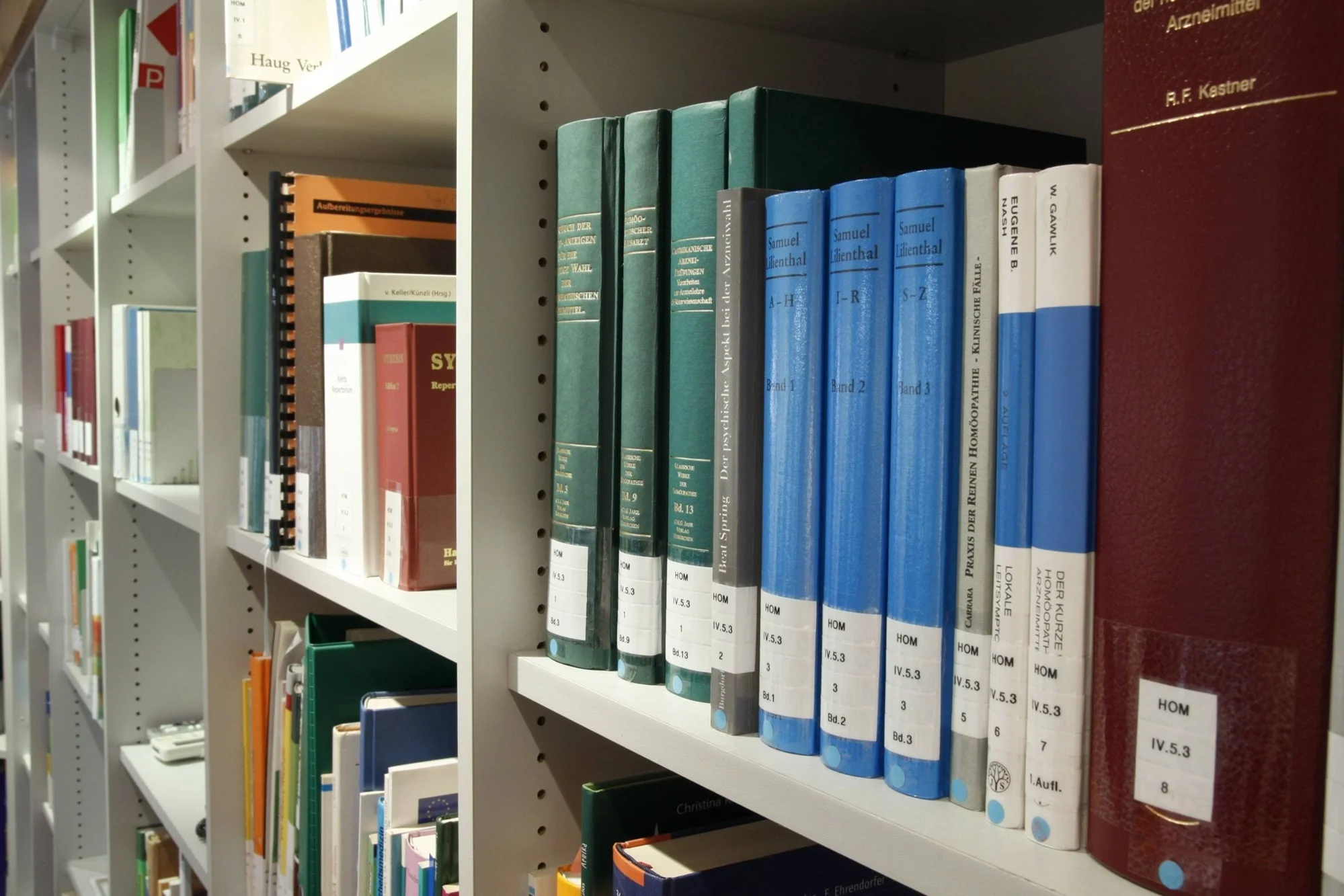 Estantes de biblioteca con libros especializados en homeopatía, clasificados con signaturas HOM, en alemán e inglés.