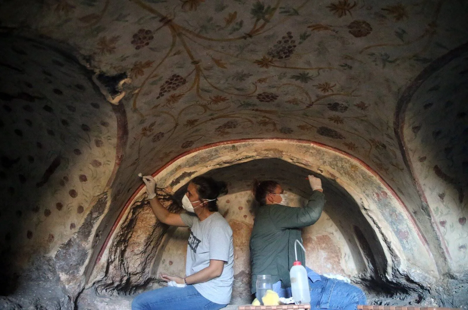 Dos restauradores con mascarilla limpian y consolidan frescos florales en la bóveda de una cámara arqueológica subterránea.