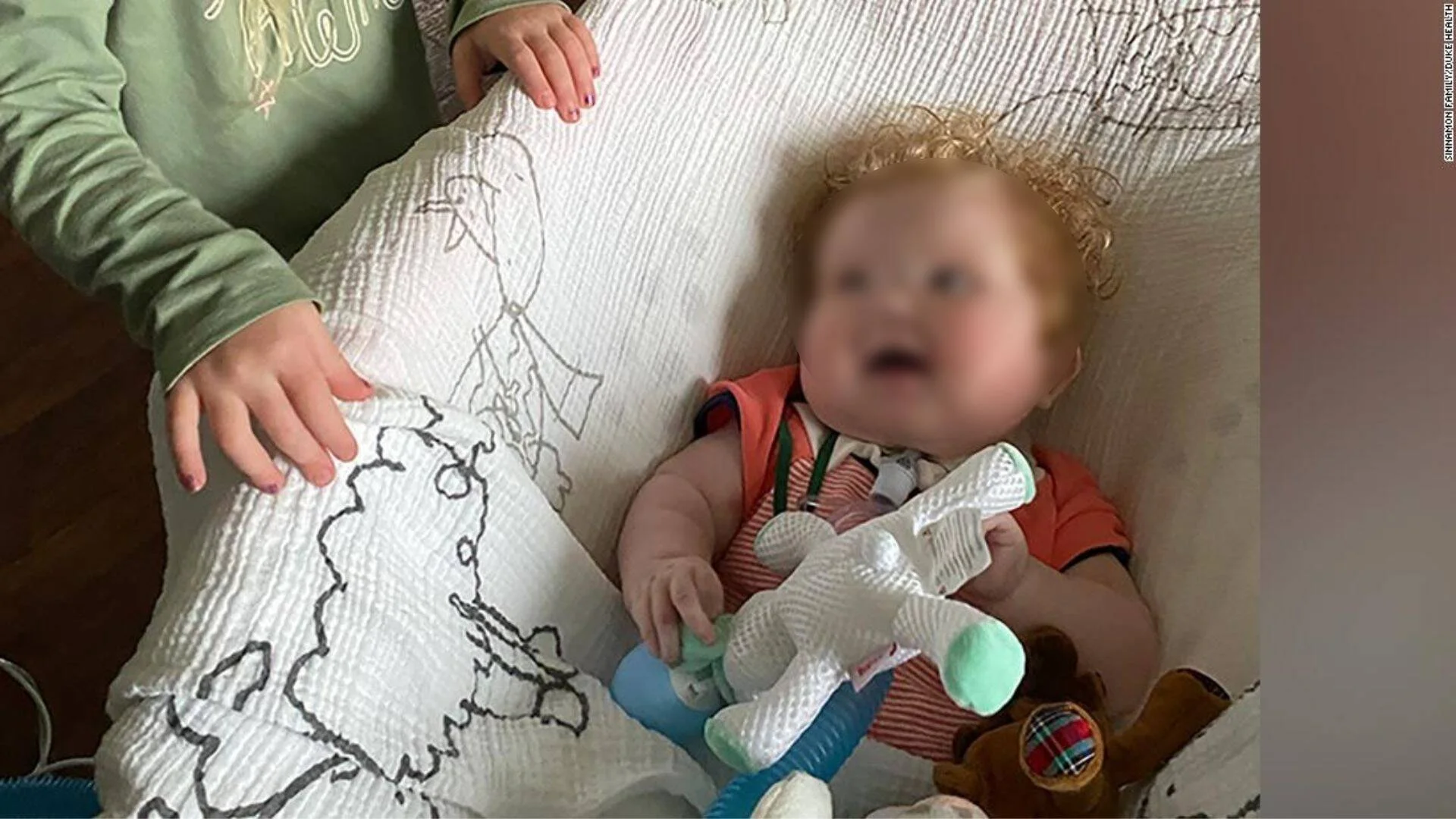 Bebé con el rostro pixelado recostado junto a juguetes y una manta con dibujos, acompañado por un niño mayor.