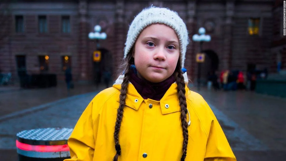 Adolescente con trenzas, gorro blanco e impermeable amarillo mira a cámara frente a un edificio de piedra.