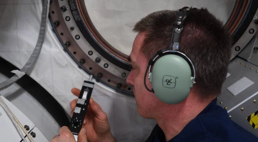 Astronauta con auriculares verdes sostiene un dispositivo electrónico junto a una escotilla de la estación espacial internacional.