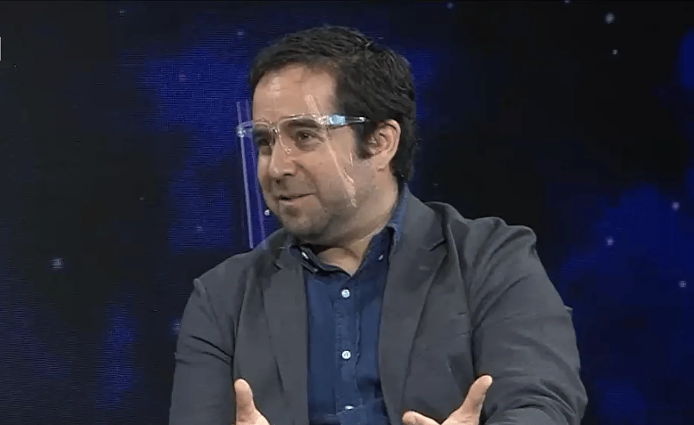 Hombre de cabello oscuro con protector facial transparente y saco gris gesticula mientras habla en un estudio de televisión.