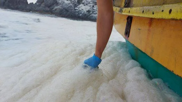 Pie calzado con escarpín azul parado en espuma de mar junto al casco de un bote de madera amarillo.