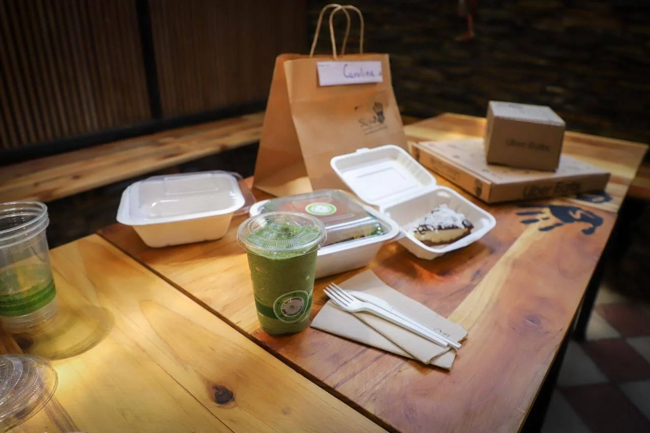 Mesa de madera con envases de comida para llevar, una bebida fría verde, cubiertos desechables y una bolsa de papel kraft con nombre escrito.