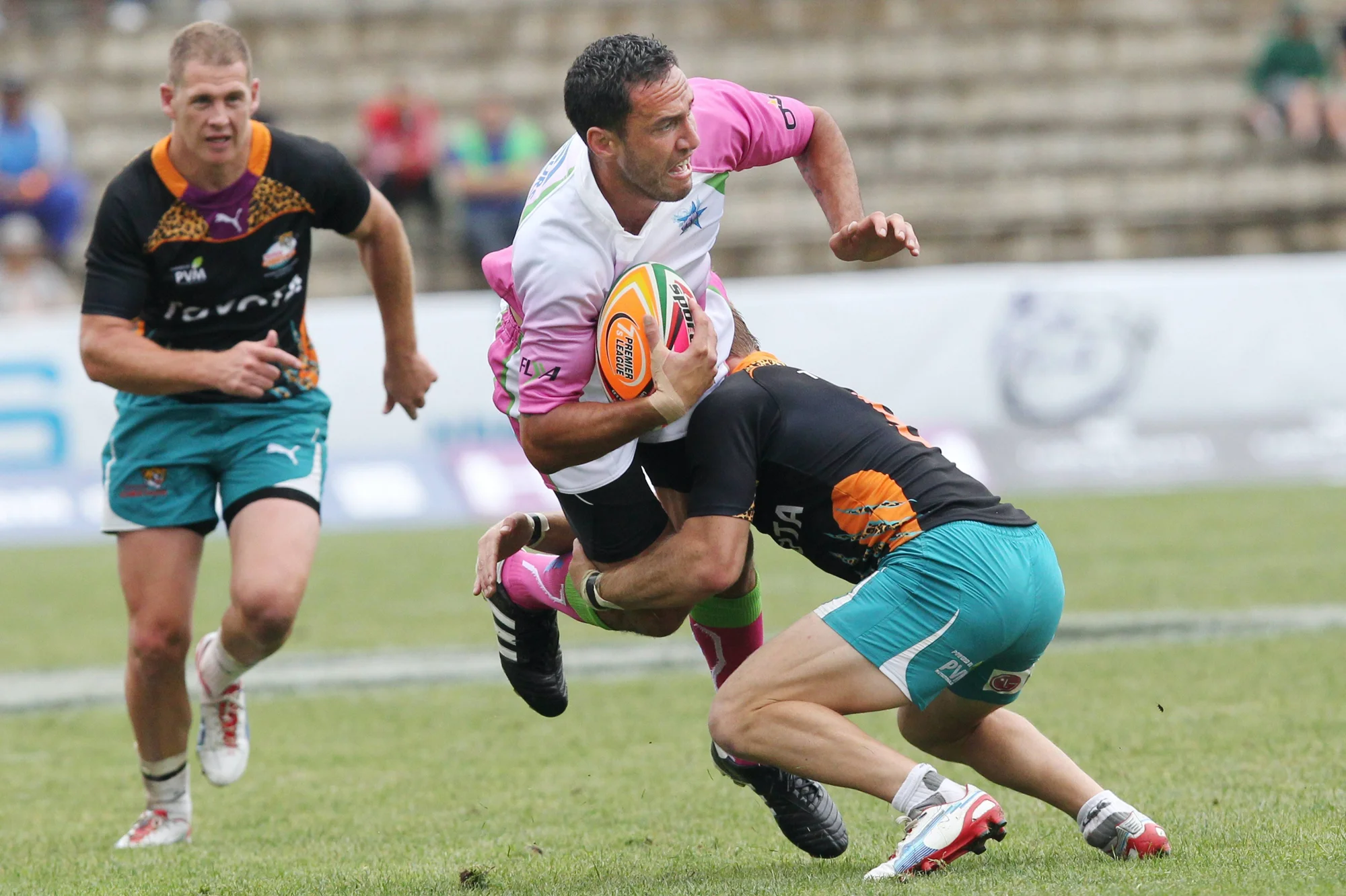 Jugador de rugby con camiseta rosa y blanca es tackleado por un rival mientras avanza con el balón en un estadio.