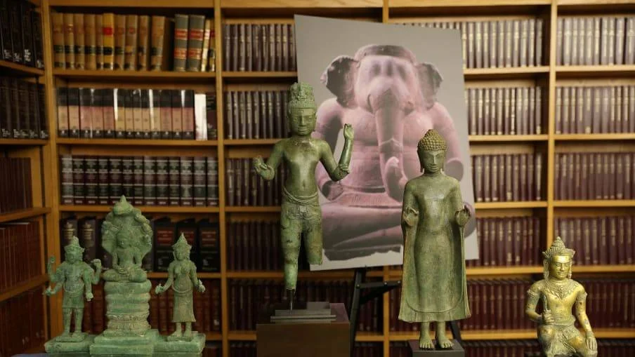 Varias estatuas de bronce de deidades budistas e hindúes exhibidas frente a estanterías con libros, junto a una fotografía de una figura de Ganesha.