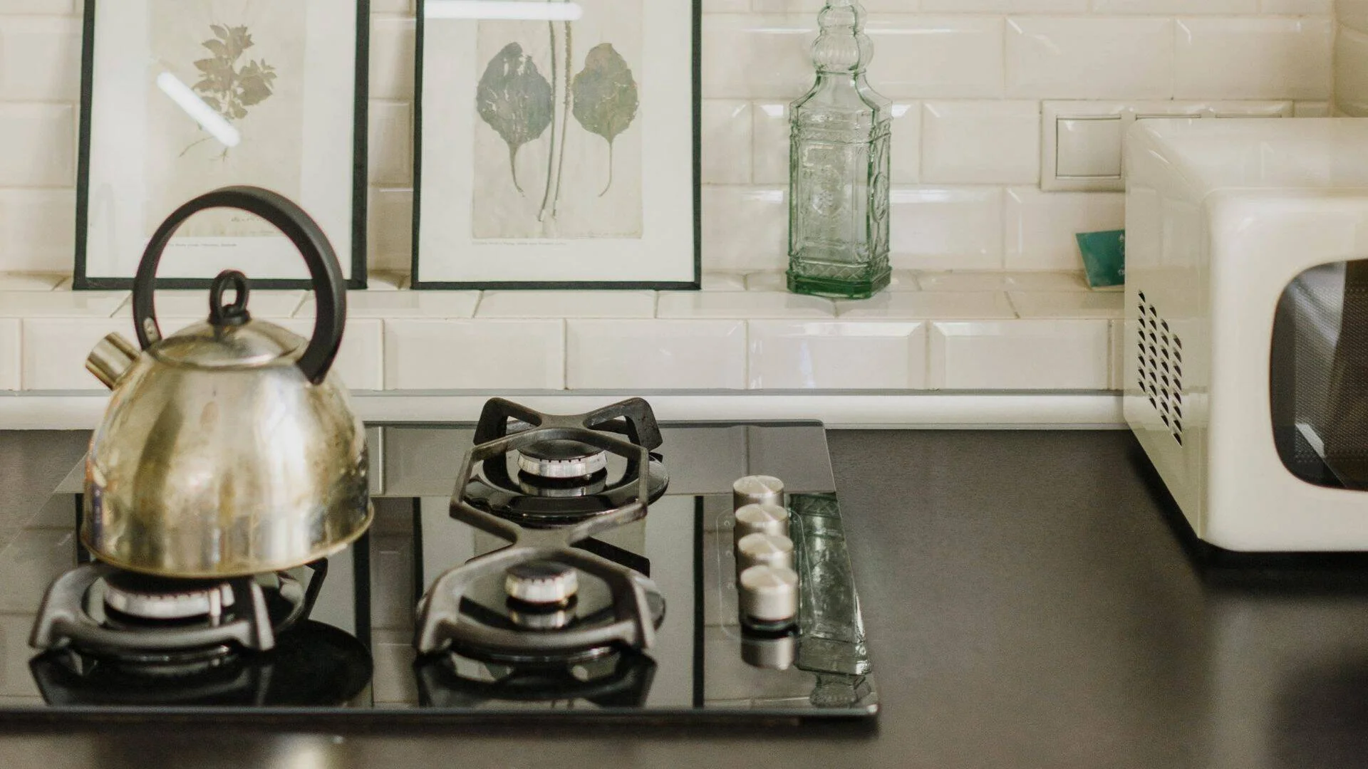 Hornalla a gas con pava metálica, microondas blanco y láminas botánicas decorativas sobre azulejos blancos.