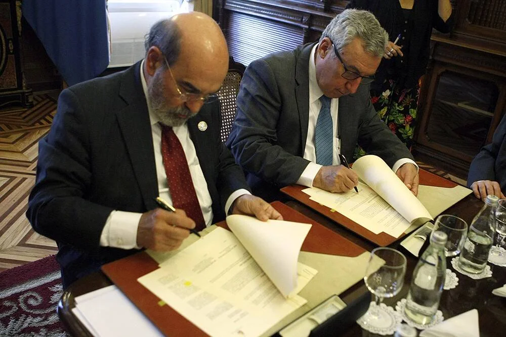 Dos hombres con traje firman documentos oficiales sentados en una mesa de reuniones.