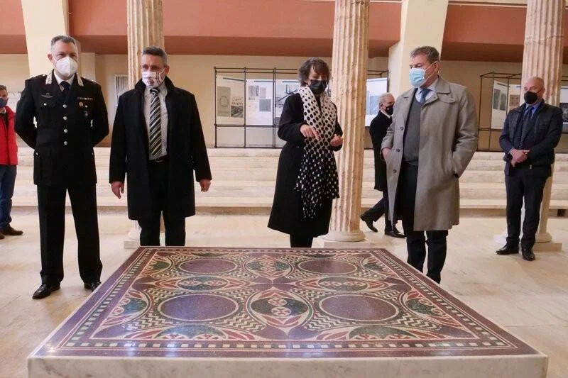 Funcionarios y oficial de carabineros con mascarilla observan un gran mosaico romano policromado en la sala de un museo.
