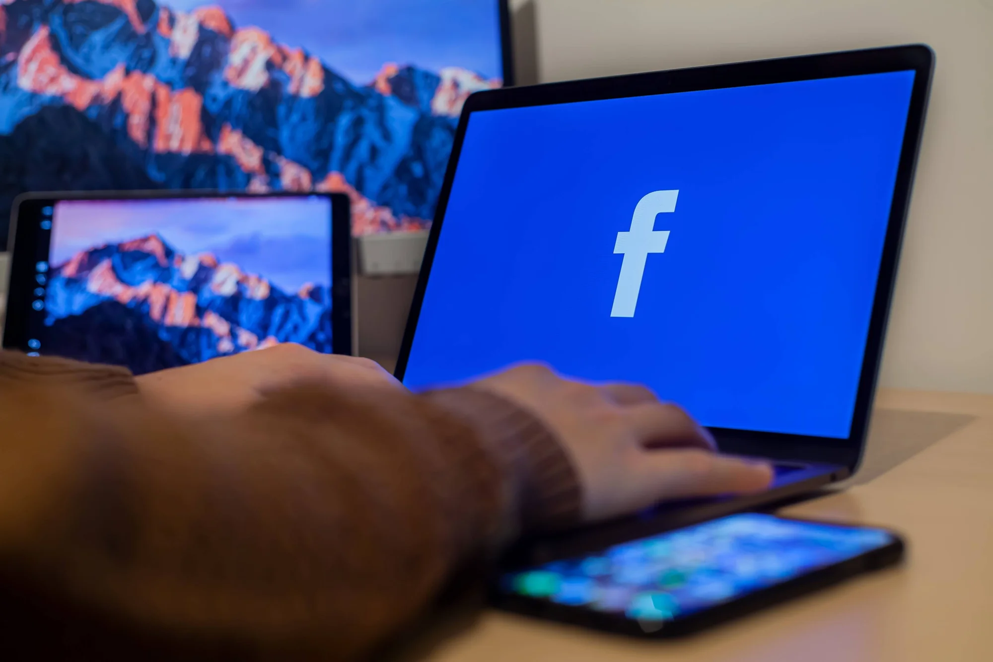 Mano sobre el teclado de una laptop con el logo de Facebook en pantalla, junto a una tablet y un smartphone.