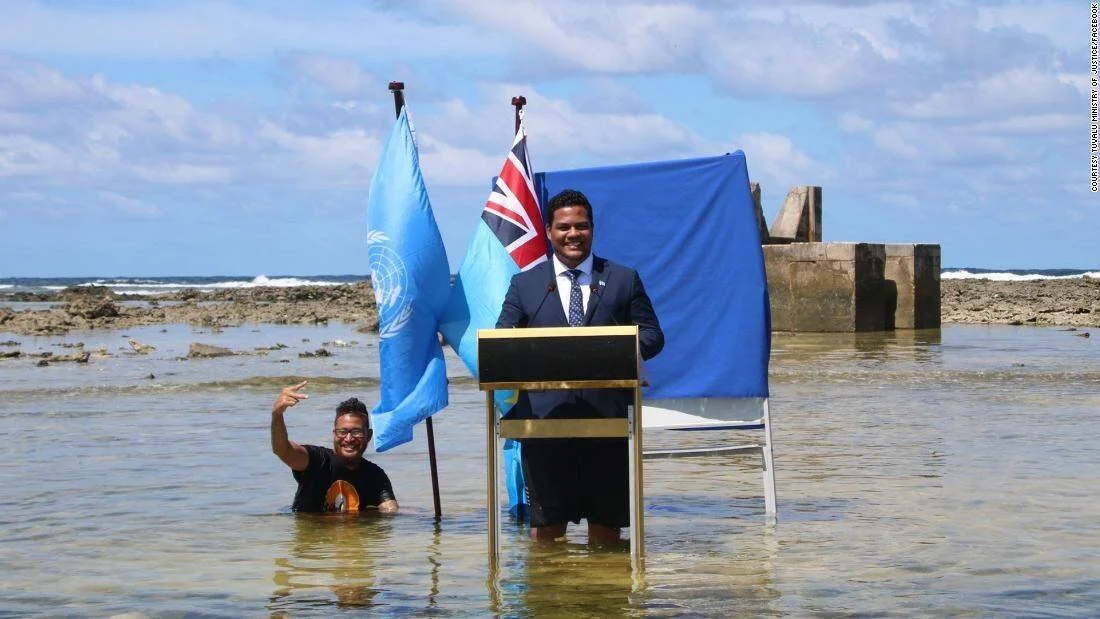 Un ministro de Tuvalu da un discurso desde un atril dentro del mar, flanqueado por la bandera de la ONU y la de Tuvalu, visibilizando el impacto del cambio climático.