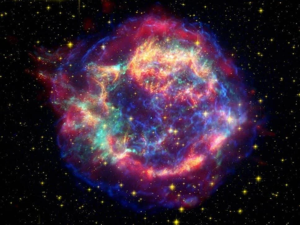 Nube esférica de gas y polvo en tonos azules, rojos y naranja producto de una explosión de supernova, rodeada de estrellas.