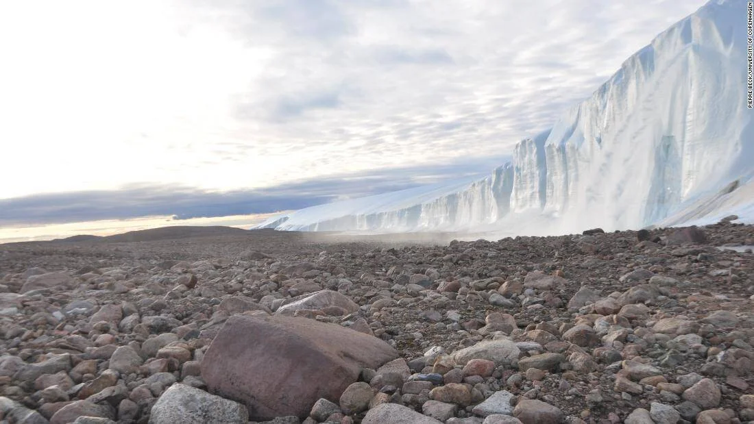 Gran pared de hielo glaciar se eleva sobre un terreno pedregoso ártico bajo un cielo nublado.