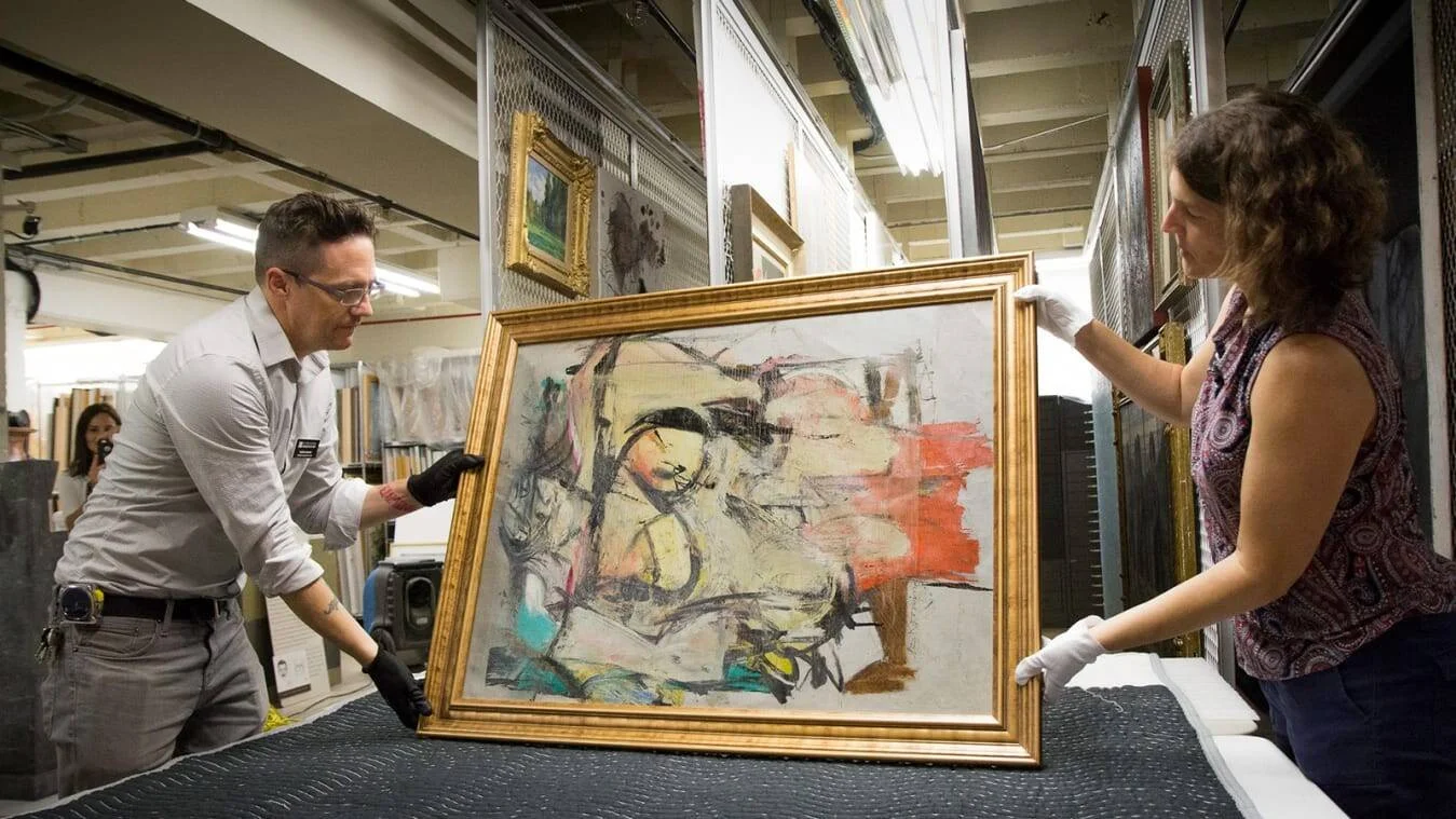 Dos personas con guantes sostienen una pintura abstracta enmarcada en oro dentro del depósito de un museo.