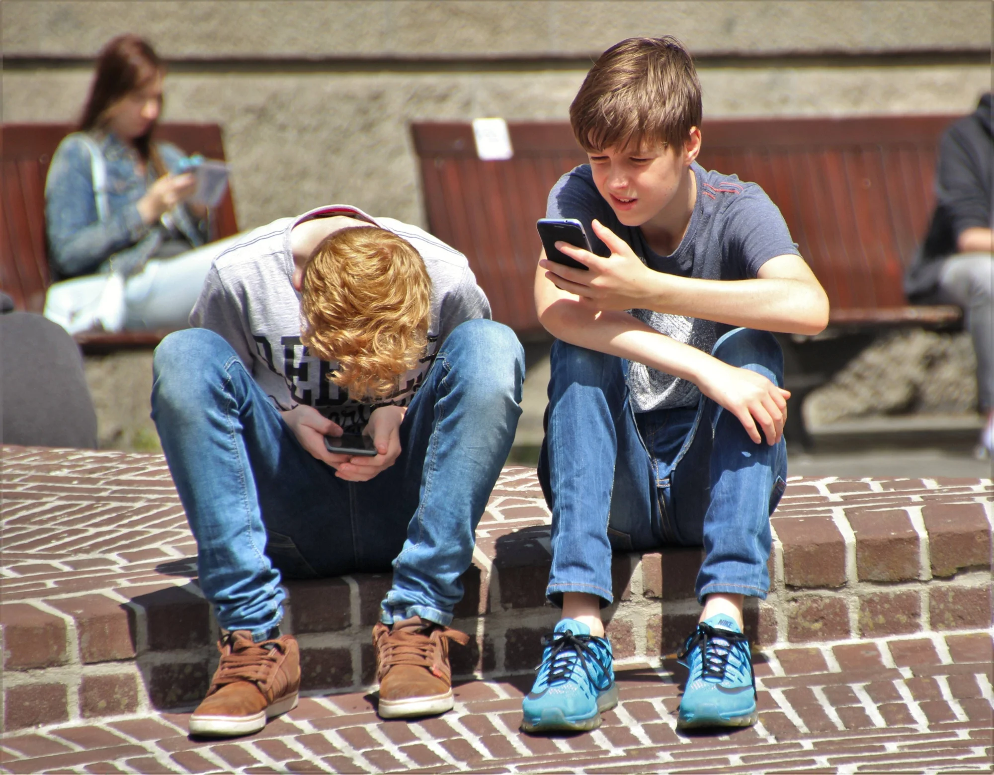 Dos chicos adolescentes sentados en un muro de ladrillos mirando sus teléfonos inteligentes en un espacio público.