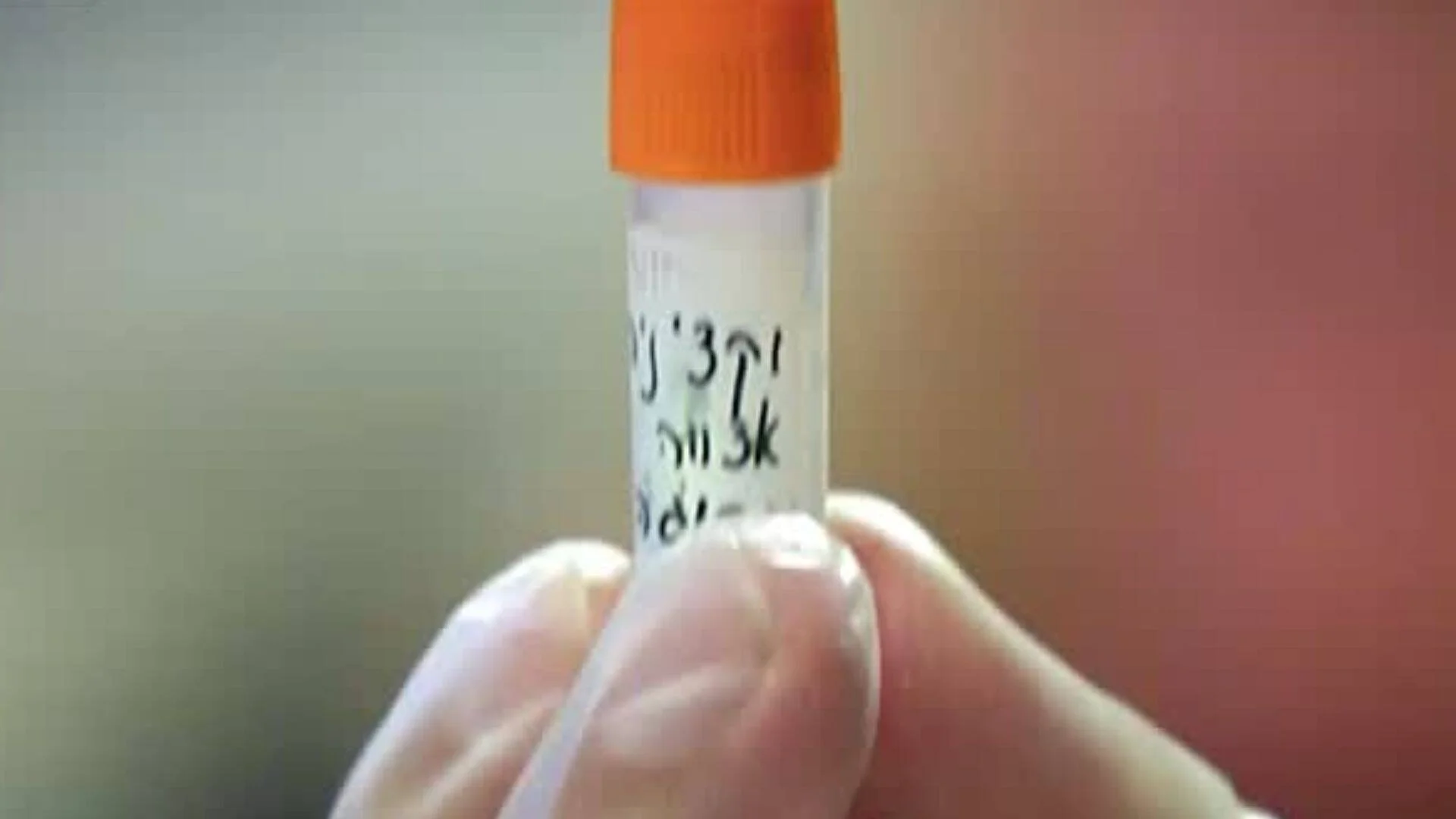 Mano con guante sostiene un tubo de muestra de laboratorio con tapa naranja y texto escrito a mano.