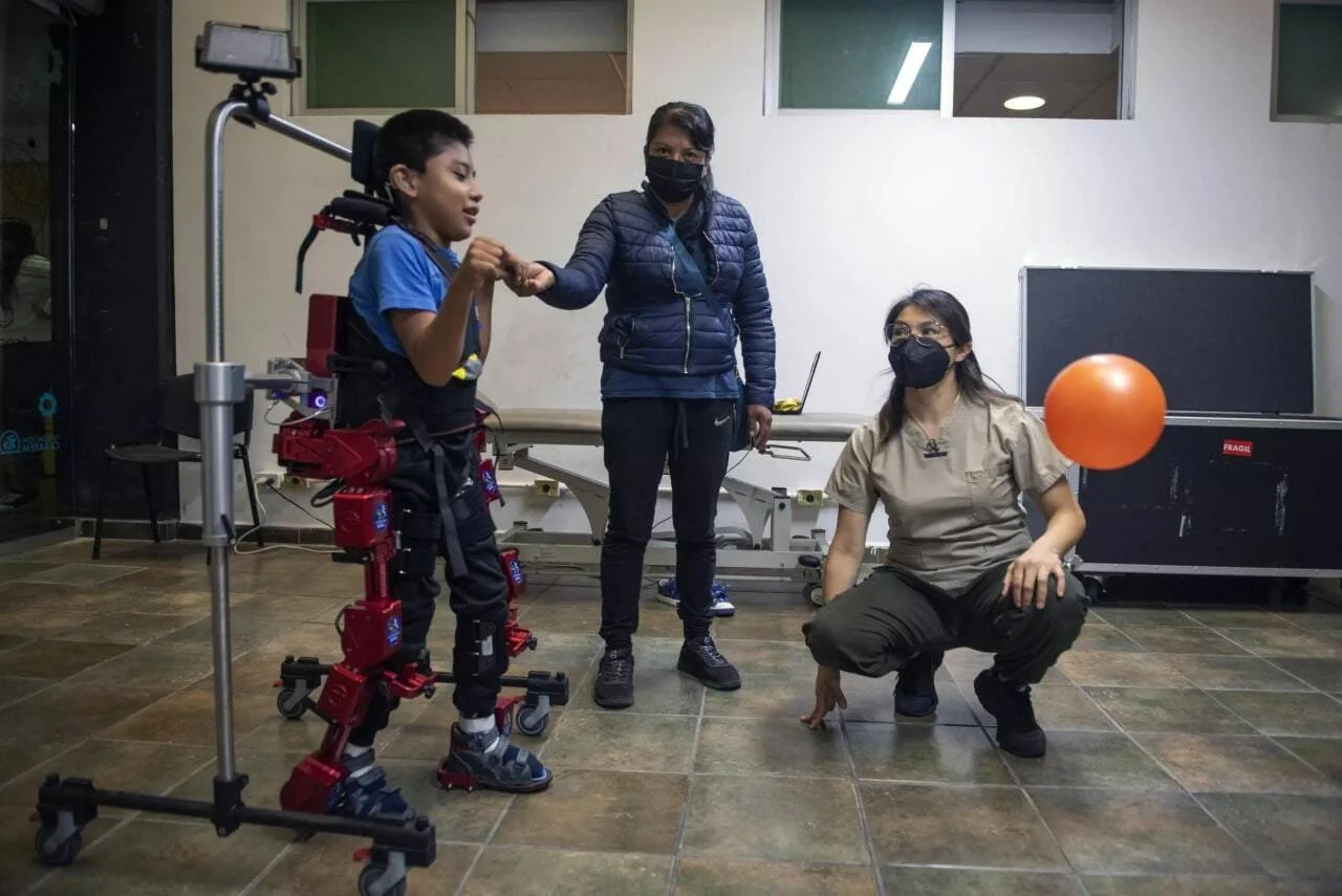 Un niño usa un exoesqueleto robótico rojo durante una sesión de rehabilitación, asistido por dos profesionales de la salud con mascarilla.
